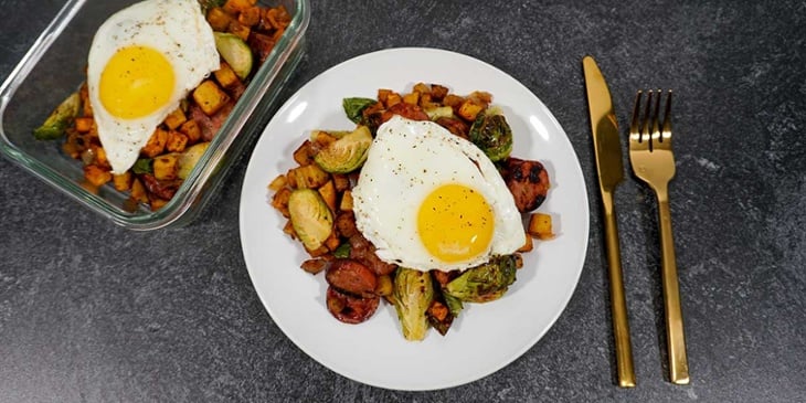 Paleo Sweet Potato Hash with Sausage Egg plated on a white plate and meal prep container on black background next to golden silverware